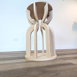May include: A light-colored wooden stool with a round top and base. The stool's design features arched legs that connect the base to the top, creating a unique, open structure. The wood has a natural, unfinished appearance.