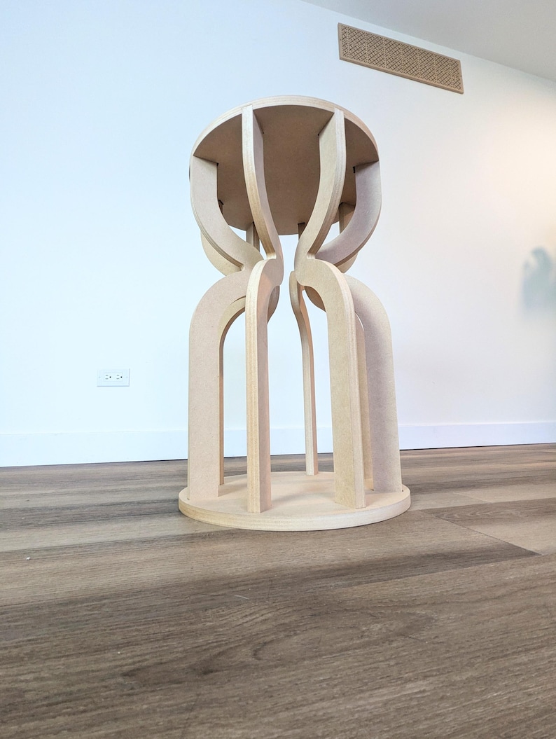 May include: A light-colored wooden stool with a round top and base. The stool's design features arched legs that connect the base to the top, creating a unique, open structure. The wood has a natural, unfinished appearance.