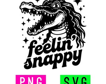 May include: A black and white graphic illustration of a smiling alligator with long hair, surrounded by small stars. Below the alligator, the text reads "feelin' snappy" in a bold, cursive font. Two rectangular boxes appear below, one pink with "PNG" and one green with "SVG".