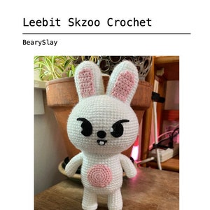 May include: White crocheted bunny with pink ears and a pink circle on its chest. The bunny has black eyes and a black mouth. The bunny is sitting on a wooden surface.