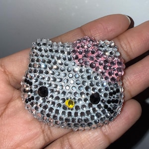 May include: A sparkling, rhinestone-covered Hello Kitty face. The iconic cat's face is covered in silver rhinestones, with a pink bow, black eyes, and a yellow nose. The item is a decorative accessory.