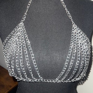 May include: Silver chainmail bra with a halter neck design. The bra is made of multiple strands of small, shiny silver chains, creating a draped effect. The chains are connected to form a triangle shape at the bust.