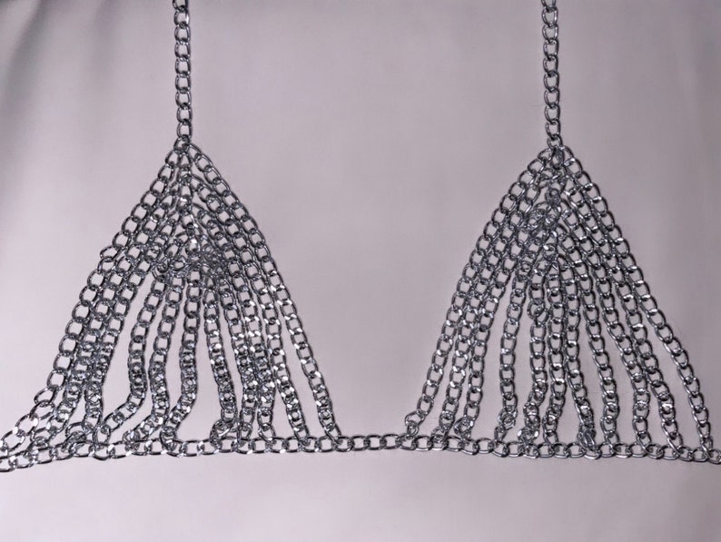 May include: A silver-colored chainmail bra with triangular cups and multiple dangling chains. The bra is made of small, interlocking metal links and has straps that go over the shoulders.