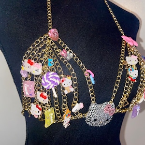 May include: Gold-coloured chain bra decorated with Hello Kitty charms, candy-themed embellishments, and a rhinestone-studded Hello Kitty face. The bra features multiple strands and colourful accents.