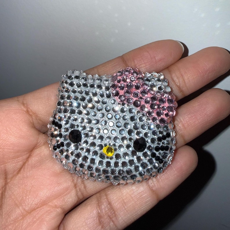 May include: A sparkling, rhinestone-covered Hello Kitty face. The iconic cat's face is covered in silver rhinestones, with a pink bow, black eyes, and a yellow nose. The item is a decorative accessory.