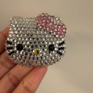 May include: A sparkly, rhinestone-covered Hello Kitty face. The face is silver with a pink bow, black eyes, and a yellow nose. The image is of a decorative item, possibly a brooch or embellishment.