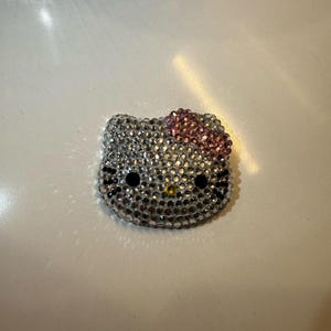 May include: A sparkling, rhinestone-covered Hello Kitty face. The cat's face is silver with black eyes, whiskers, and a yellow nose. A pink bow sits atop the head. The item is likely a decorative accessory or embellishment.