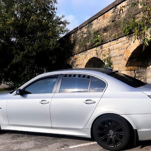 May include: Silver saloon car with black alloy wheels and a rear spoiler parked on a paved area. The car has tinted windows and a sunroof. The background features a stone wall with arches and a tree.