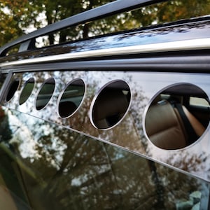 May include: A black car with a tinted window that has a row of round holes cut out of it. The holes are evenly spaced and are about the size of a 20 pence coin. The window is reflecting the trees outside.