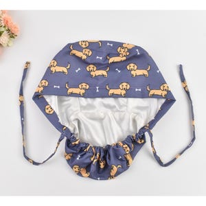 May include: A blue surgical scrub cap featuring a repeating pattern of cartoon dachshund dogs and white dog bones. The cap has a white satin lining and long blue ties for securing. The cap is designed to cover the hair.