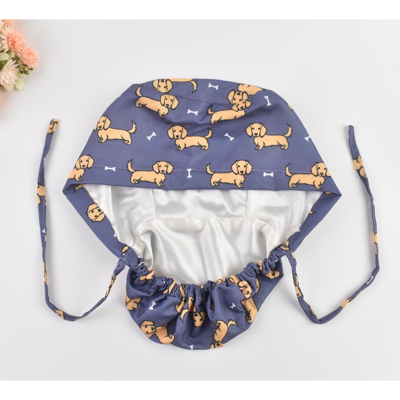 May include: A blue surgical scrub cap featuring a repeating pattern of cartoon dachshund dogs and white dog bones. The cap has a white satin lining and long blue ties for securing. The cap is designed to cover the hair.