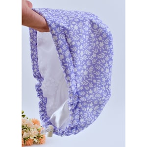 Puede incluir: Un gorro de ducha lavanda con un estampado floral blanco. El gorro tiene una banda elástica y un interior blanco. El gorro está sostenido por una mano, y un pequeño ramo de flores está en el fondo.