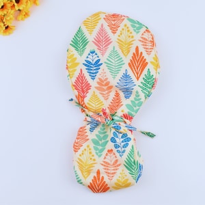 May include: A white scrub cap with a colorful leaf pattern. The leaves are in shades of blue, green, orange, and pink. The cap has a tie at the back.
