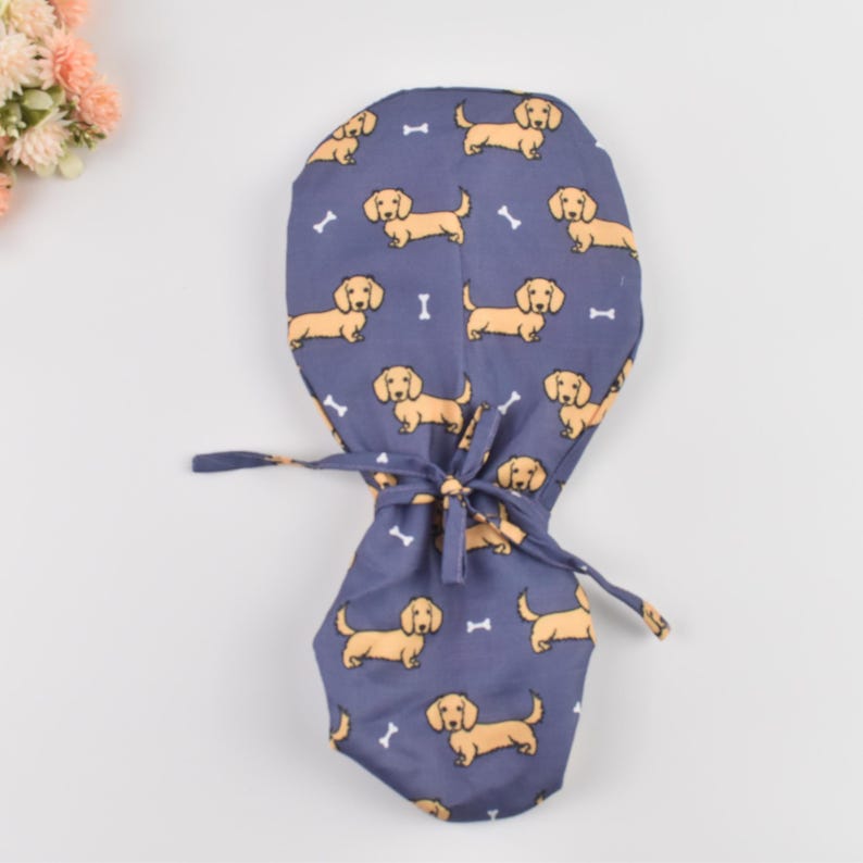 May include: A blue surgical scrub cap featuring a repeating pattern of tan dachshund dogs and white bone shapes. The cap has a tie closure and is designed for medical professionals or pet lovers.