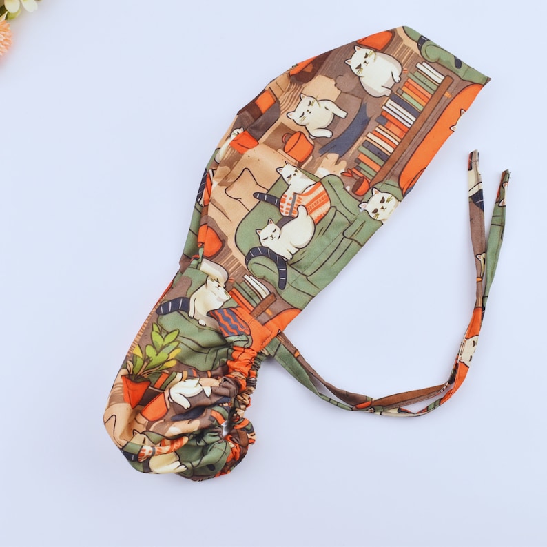 May include: A patterned scrub cap with a repeating design of white cats on green sofas and bookshelves. The cap has an orange and brown colour scheme with green accents and ties at the back for an adjustable fit.