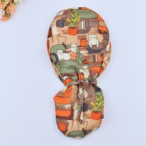 May include: A scrub cap with a repeating pattern of white cats on green sofas, bookshelves, and plants. The cap's colours are brown, orange, and green. It has a tie closure at the back. The design is reminiscent of a cozy living room.