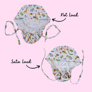 May include: Two surgical scrub caps, one not lined and one satin lined, are displayed. The caps feature a white background with a colorful safari animal print, including giraffes, elephants, and zebras. The caps have ties for a secure fit.