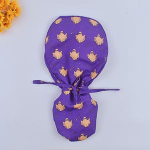 May include: A purple scrub cap with a Halloween ghost pattern. The cap features a repeating pattern of light orange ghosts and bone shapes. The cap has a tie closure at the back.