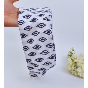 May include: White fabric with a repeating pattern of navy blue diamond shapes. The fabric is held in a hand, and a small bouquet of white flowers is in the background.