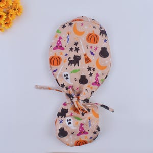 May include: A tan scrub hat with a Halloween-themed print featuring black cats, pumpkins, witches hats, ghosts, and stars.