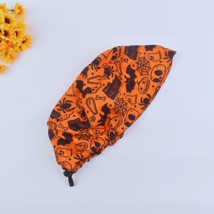 May include: Orange Halloween-themed scrub cap with a black pattern of bats, spiders, cobwebs, skulls, and tombstones. The cap has a drawstring closure and is made of a lightweight fabric. Perfect for healthcare workers or anyone wanting to celebrate Halloween.