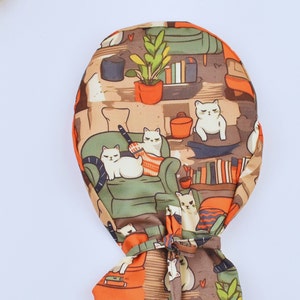 May include: A patterned fabric item featuring a repeating design of white cats in a living room scene. The design includes bookshelves, potted plants, and orange accents. The item is tied at the bottom with a matching fabric tie.