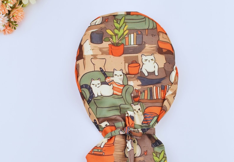 May include: A patterned fabric item featuring a repeating design of white cats in a living room scene. The design includes bookshelves, potted plants, and orange accents. The item is tied at the bottom with a matching fabric tie.