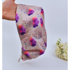 Puede incluir: Un artículo de tela beige con un colorido patrón abstracto de formas moradas, rosas y azules. El artículo se sostiene en una mano y parece ser un cubrecabezas o accesorio. El fondo es blanco.