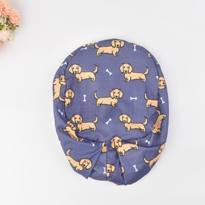 May include: A blue fabric hat with a repeating pattern of tan dachshund dogs and white bone shapes. The hat is gathered at the bottom and has a rounded shape. The background is white.