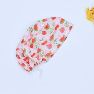 May include: Pink surgical scrub cap with a repeating pattern of red watermelon slices, strawberries, cherries, and peaches. The cap also features white flowers and green leaves. The cap has a drawstring closure.