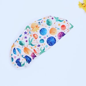 May include: A white, patterned shower cap featuring a colorful celestial design. The cap is adorned with planets, stars, and constellations in shades of blue, orange, purple, and green. The cap is designed to protect hair from water.