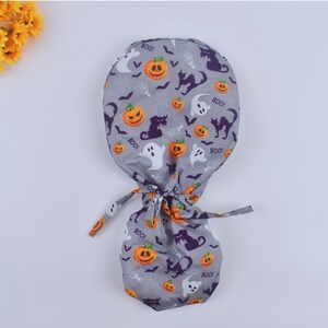 May include: Gray scrub hat with a Halloween print featuring pumpkins, ghosts, bats, and black cats. The hat has a tie at the back.