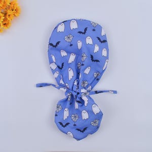 May include: A blue scrub cap featuring a Halloween-themed pattern. The cap is adorned with white ghosts, black bats, and spiderwebs. It has a tie closure at the back. The cap is designed for medical professionals or those in the healthcare field.