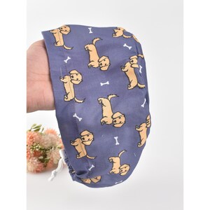 May include: A blue surgical scrub cap featuring a repeating pattern of tan-colored dogs and white bone shapes. The cap is held in a hand and has a white drawstring.