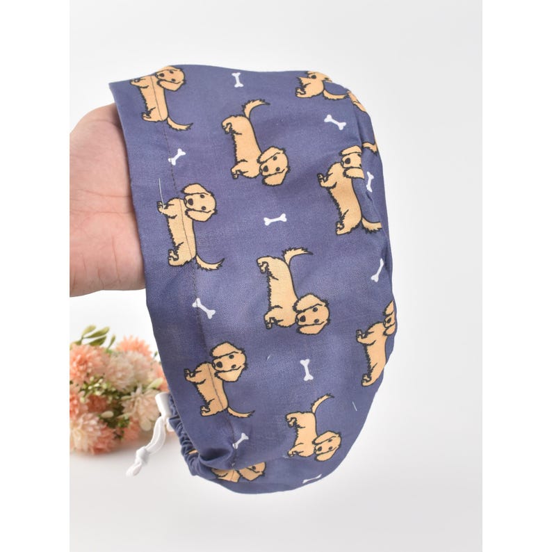 May include: A blue surgical scrub cap featuring a repeating pattern of tan-colored dogs and white bone shapes. The cap is held in a hand and has a white drawstring.
