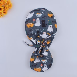May include: A gray and white surgical scrub hat with a Halloween theme. The hat features a variety of Halloween characters, including ghosts, pumpkins, bats, and a black cat. The hat has a tie at the back.
