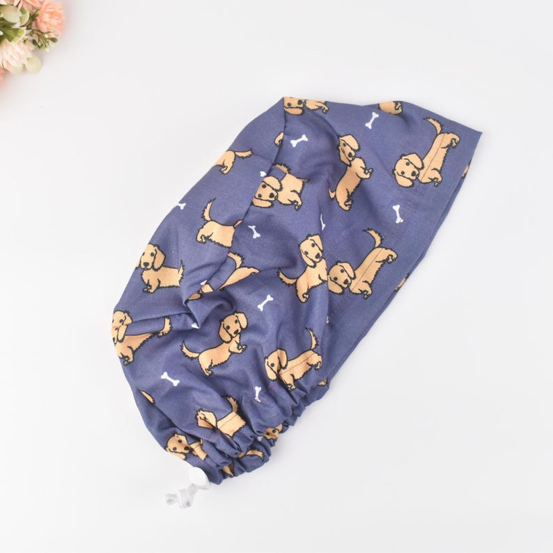 May include: A blue fabric item with a repeating pattern of dachshund dogs and white bones. The fabric is gathered at the bottom with a white drawstring. The item is likely a shower cap or similar head covering.