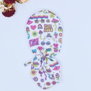 May include: White scrub cap with a colorful, whimsical design. The cap features various illustrations including mushrooms, rainbows, hearts, and the words "Good Vibes" and "Peace Love". The design is in shades of pink, yellow, blue, and green.