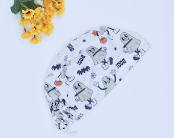 May include: White Halloween-themed scrub cap with a repeating pattern of cartoon ghosts, bats, and spiderwebs. The ghosts are gray with black eyes and are holding pumpkins. The word "BOO!" is printed on the cap.
