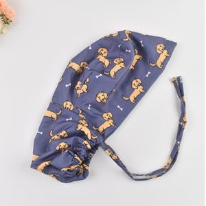 May include: A navy blue scrub cap featuring a repeating pattern of tan dachshund dogs and white bone shapes. The cap has a gathered back and long ties for securing. The fabric is a soft, lightweight material.