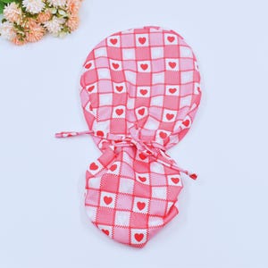 May include: A pink and white patterned scrub cap with a red heart design. The cap is tied at the back with matching fabric ties. The fabric has a checkerboard pattern with red hearts in each square.