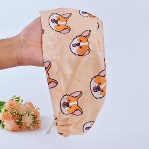 Può includere: Un cappello da scrub color pesca con un motivo ripetuto di corgi dei cartoni animati. I corgi sono marroni, bianchi e arancioni con nasi neri e occhi neri.