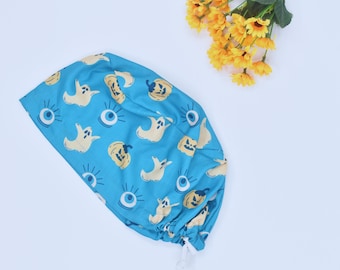 May include: A blue Halloween-themed scrub cap featuring a repeating pattern of cartoon ghosts, jack-o'-lanterns, and eyeballs. The cap has a drawstring closure and is made of a lightweight fabric. Yellow flowers are in the background.