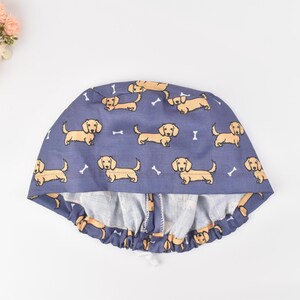 May include: A blue surgical scrub cap featuring a repeating pattern of tan dachshund dogs and white bone shapes. The cap has a gathered elastic band and a white drawstring for a secure fit. The fabric is a soft, lightweight material.