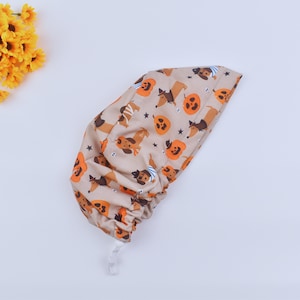 May include: A tan scrub hat with a Halloween-themed print featuring orange pumpkins and brown dachshunds wearing costumes. The hat has a white drawstring.