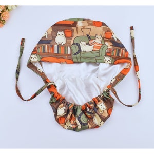 May include: A patterned scrub cap with a cat design. The cap's exterior features cats on furniture and bookshelves in brown, orange, and green. The interior is a smooth, white fabric, and it has tie closures. A practical and stylish accessory.