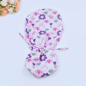 May include: A pink scrub cap with a repeating pattern of purple flowers, stars, hearts, rainbows, and green leaves. The cap is tied at the back with two long ties. The cap is designed for medical professionals.