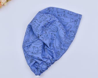 May include: A blue surgical scrub cap with a repeating pattern of black cat illustrations. The cap has a drawstring for adjustment. The fabric is soft and the design is playful, suitable for medical staff or cat enthusiasts. The cap is approximately 25 cm in diameter.