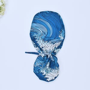 Può includere: Un cappuccio per scrub a fantasia blu e bianca con un motivo a onde. Il cappuccio è legato sul retro con un laccio in tessuto abbinato. Il motivo a onde include sfumature di blu e bianco, creando un design dinamico e visivamente accattivante.