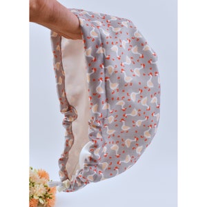 Könnte beinhalten: Ein graues Stoffstück mit einem sich wiederholenden Muster aus weißen und pfirsichfarbenen Gänsen mit orangefarbenen Füßen und Schnäbeln. Das Innenfutter ist hellpfirsichfarben. Der Artikel wird offen gehalten und gibt den Blick ins Innere frei. Ein weißer Kordelzug ist sichtbar.